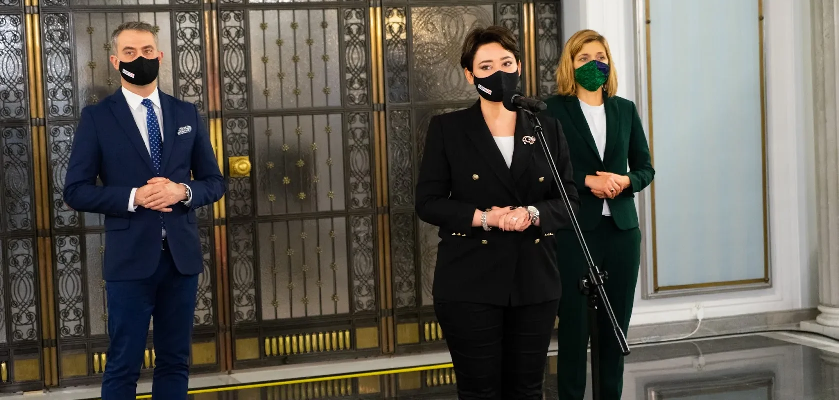 Trzy osoby w eleganckich garniturach i żakietach stoją w holu z ozdobnymi metalowymi drzwiami, wszystkie mają maseczki, a kobieta w środku stoi przy mikrofonie.