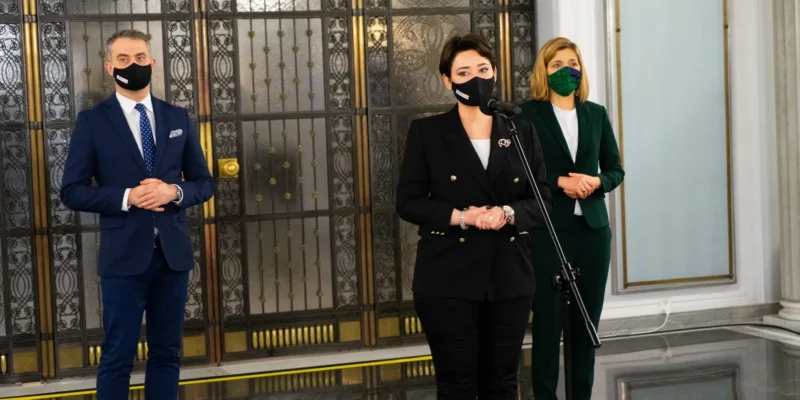 Trzy osoby w eleganckich garniturach i żakietach stoją w holu z ozdobnymi metalowymi drzwiami, wszystkie mają maseczki, a kobieta w środku stoi przy mikrofonie.