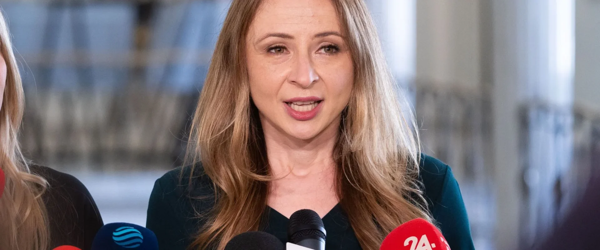 Kobieta przemawia do mediów podczas konferencji prasowej, otoczona mikrofonami.