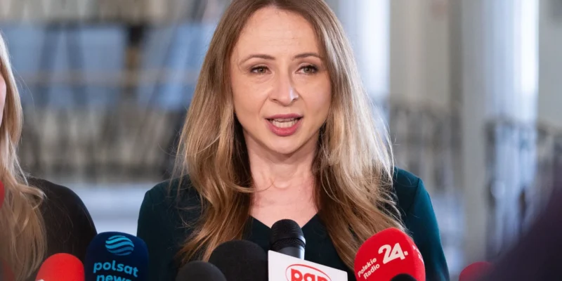 Kobieta przemawia do mediów podczas konferencji prasowej, otoczona mikrofonami.