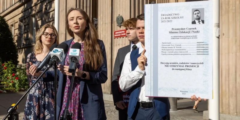 Grupa osób na zewnątrz budynku podczas konferencji prasowej; kobieta w kwiecistej sukience przemawia do mikrofonów, a obok niej ktoś trzyma duży plakat z napisem „ŚWIADECTWO ZA ROK SZKOLNY 2021/2022”.