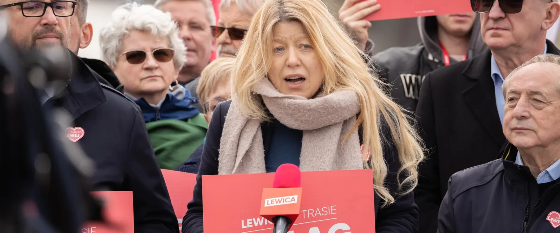 Kobieta z długimi blond włosami, owinięta szalem, przemawia do mikrofonu podczas demonstracji Lewicy, trzyma czerwony plakat, a wokół niej stoi tłum protestujących z podobnymi znakami.