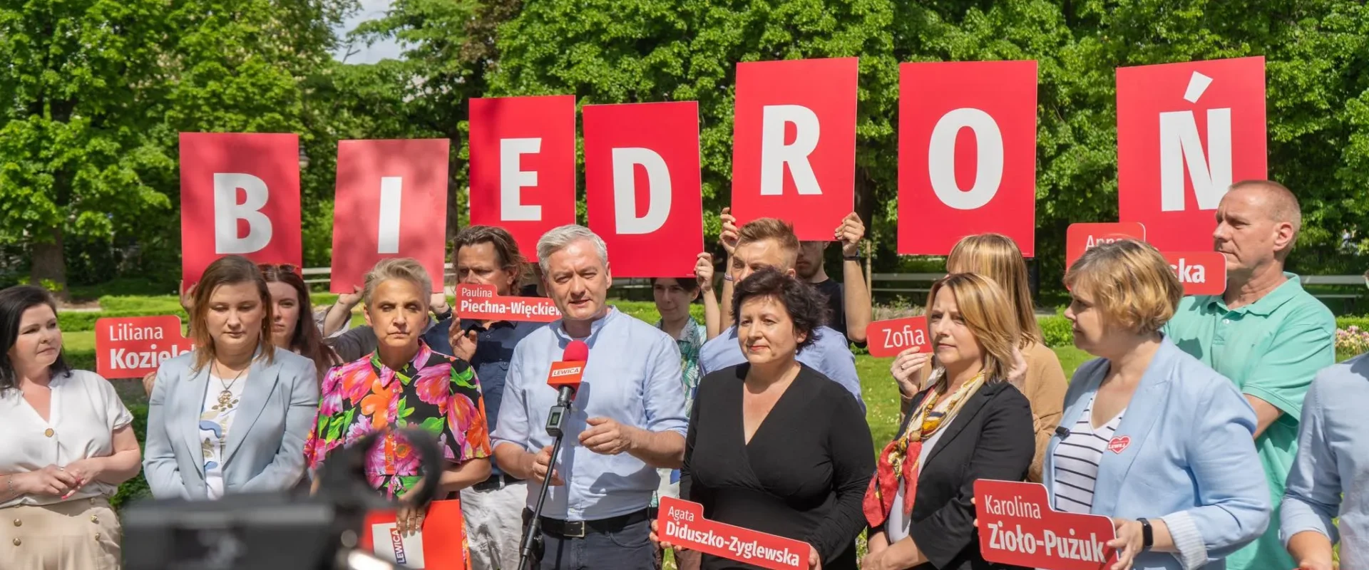 Grupa polityków stojąca na zewnątrz, trzymająca czerwone tablice tworzące napis BIEDRÓŃ, podczas gdy mężczyzna przemawia do mikrofonu.