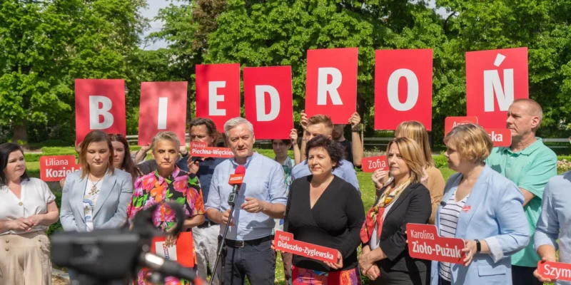 Grupa polityków stojąca na zewnątrz, trzymająca czerwone tablice tworzące napis BIEDRÓŃ, podczas gdy mężczyzna przemawia do mikrofonu.