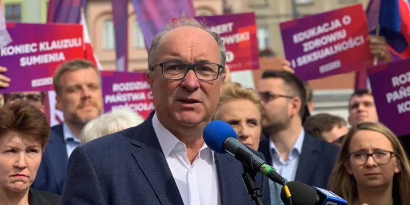 Na zdjęciu widać mężczyznę przemawiającego do mikrofonu, w otoczeniu protestujących osób trzymających kolorowe plakaty z hasłami.