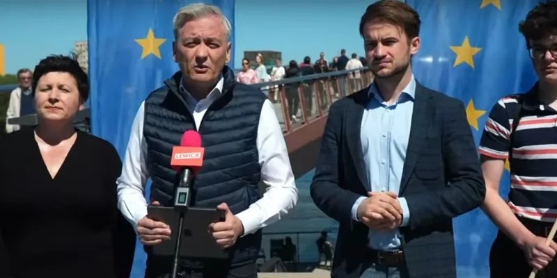 Grupa osób podczas konferencji prasowej stojąca przed niebieskim tłem z gwiazdami UE, z mówcą trzymającym różowy mikrofon z napisem LEWICA, w kontekście Europejskiej Biblioteki Cyfrowej.