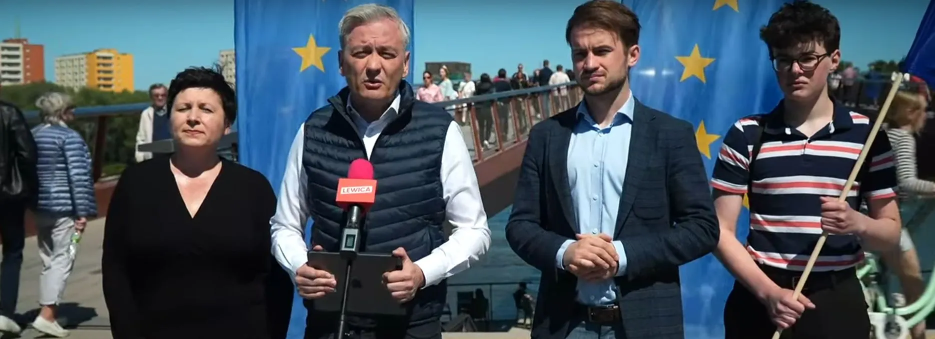 Grupa osób podczas konferencji prasowej stojąca przed niebieskim tłem z gwiazdami UE, z mówcą trzymającym różowy mikrofon z napisem LEWICA, w kontekście Europejskiej Biblioteki Cyfrowej.