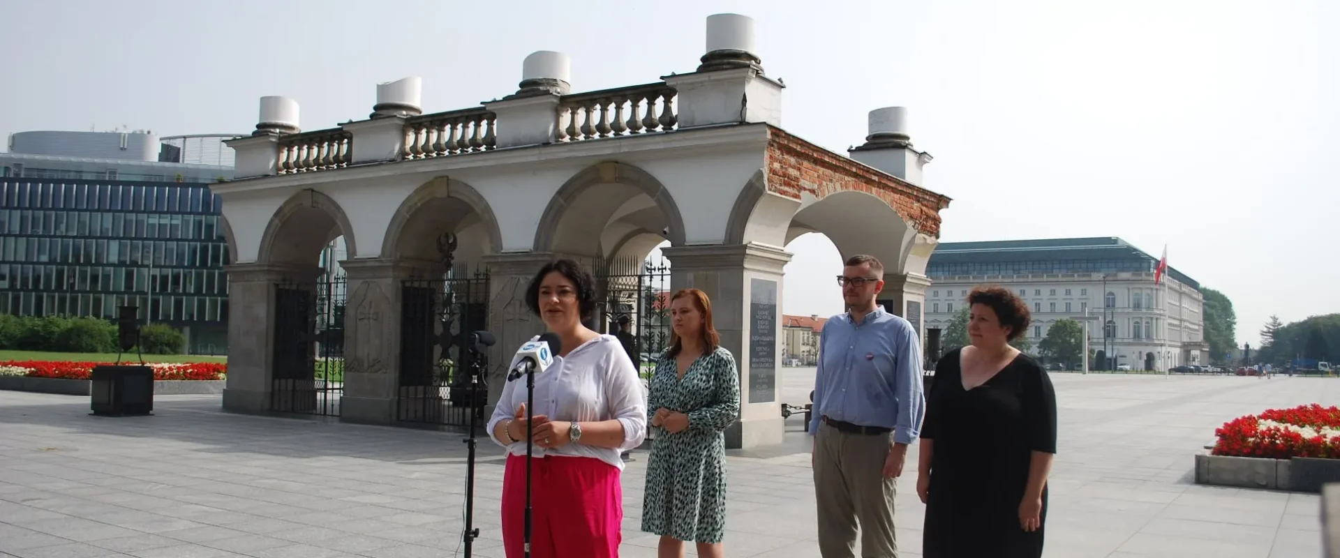Kobieta przemawia do mikrofonu podczas konferencji prasowej na placu przed zabytkowym pawilonem z arkadami; w grupie stoją inni przedstawiciele, a w tle widać nowoczesne budynki i czerwone kwiaty.