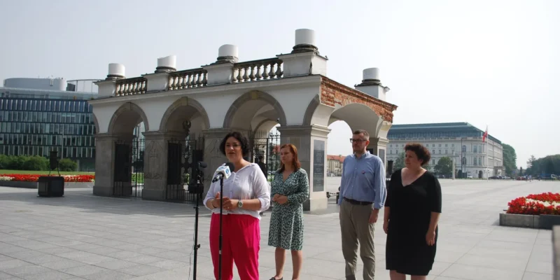 Kobieta przemawia do mikrofonu podczas konferencji prasowej na placu przed zabytkowym pawilonem z arkadami; w grupie stoją inni przedstawiciele, a w tle widać nowoczesne budynki i czerwone kwiaty.