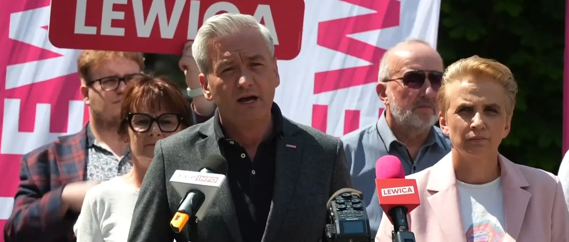 Polityk w szarej marynarce przemawia do mediów podczas konferencji prasowej, obok niego mikrofony z logiem Lewicy, a w tle widać różowy baner z napisem Lewica.