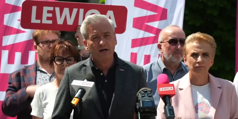 Polityk w szarej marynarce przemawia do mediów podczas konferencji prasowej, obok niego mikrofony z logiem Lewicy, a w tle widać różowy baner z napisem Lewica.