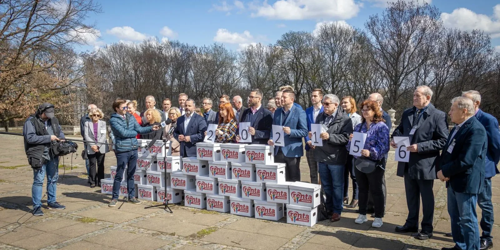 Grupa ludzi na zewnątrz prezentuje 200 000 podpisów pod obywatelskim projektem ustawy „Renta Wdowia”, stojąc obok stosów pudełek z logo projektu.