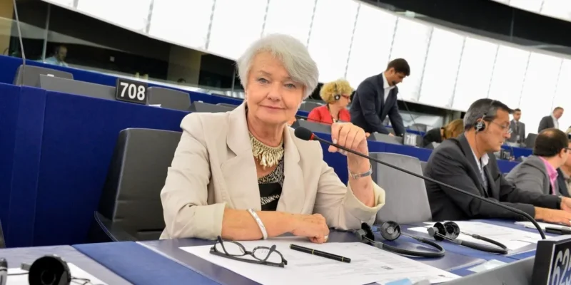 Kobieta w jasnej marynarce siedzi przy mikrofonie i dokumentach w sali obrad parlamentu.