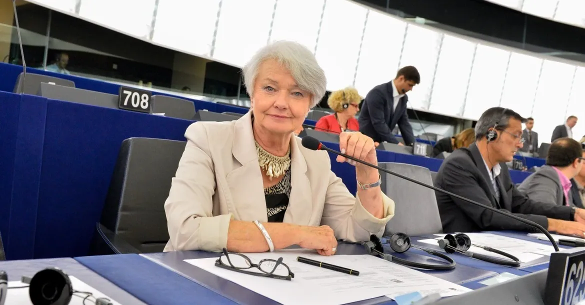 Kobieta w jasnej marynarce siedzi przy mikrofonie i dokumentach w sali obrad parlamentu.