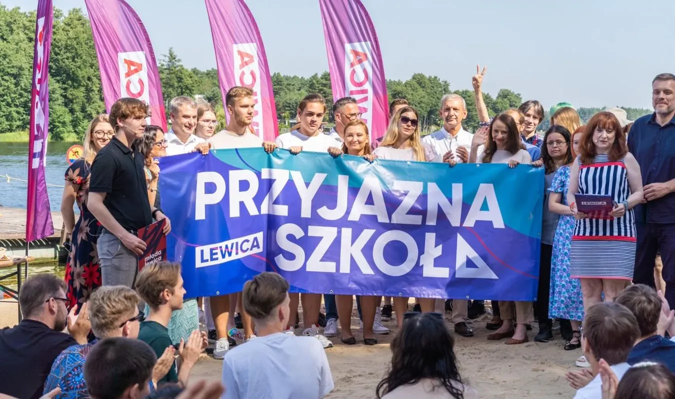 Grupa osób na plaży trzyma duży banner z napisem „PRZYJAZNA SZKOŁA” podczas konwencji programowej Lewicy, w tle widoczne fioletowe flagi i publiczność.