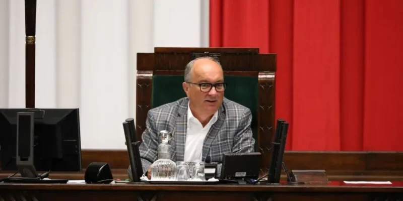 Mężczyzna w okularach i kraciastym garniturze siedzi za pulpitem w sali obrad, obok mikrofony i karafka z wodą, na tle czerwonych zasłon.