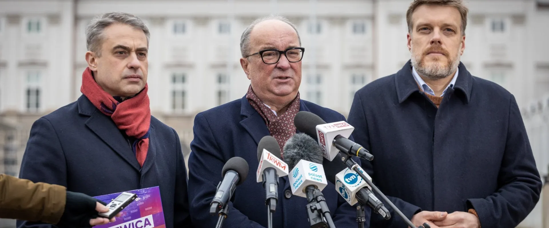 Na zdjęciu trzech mężczyzn stoi przed budynkiem, przedstawiając się jako przedstawiciele Lewicy, z mikrofonami i plakatem w dłoni.