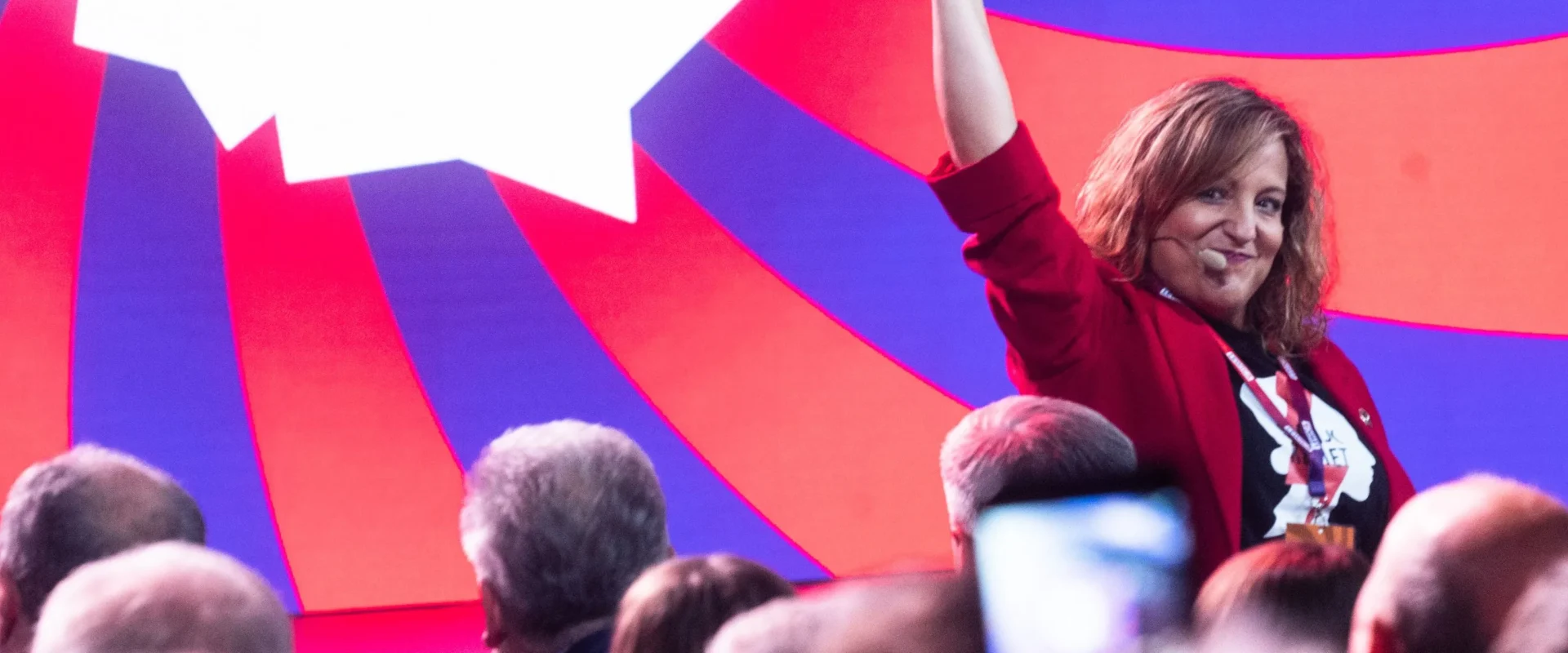 Kobieta na scenie w czerwonej marynarce z mikrofonem na uchu unosi prawą rękę w górę przed kolorowym, geometrycznym tłem i zgromadzonym tłumem widzów.