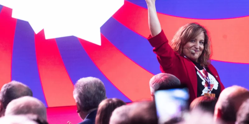 Kobieta na scenie w czerwonej marynarce z mikrofonem na uchu unosi prawą rękę w górę przed kolorowym, geometrycznym tłem i zgromadzonym tłumem widzów.