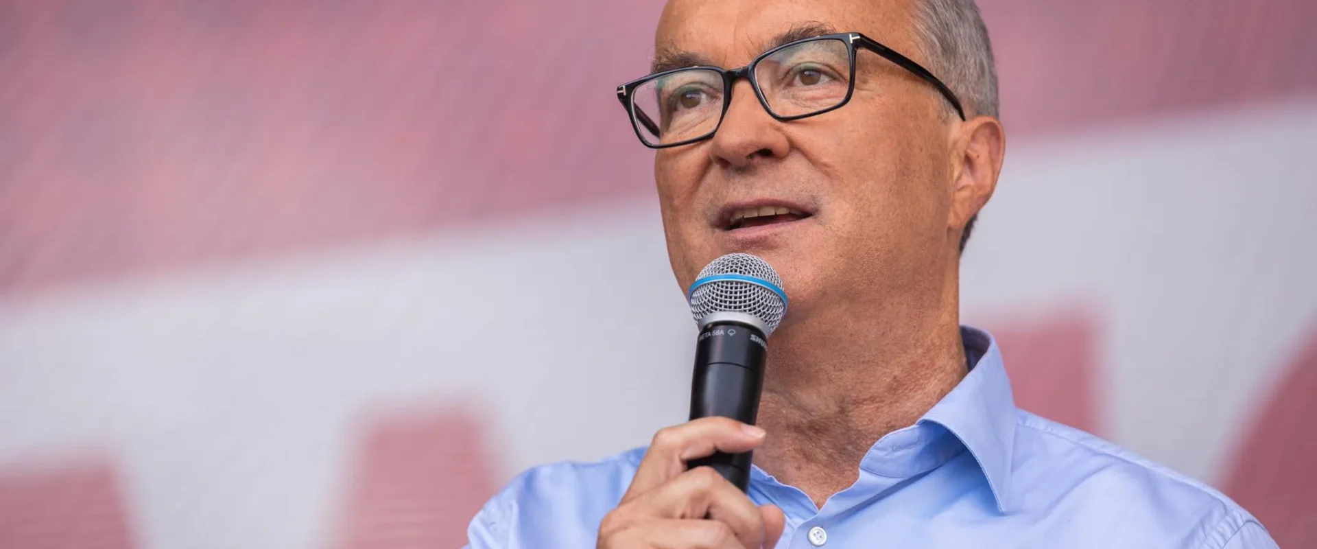 Mężczyzna w okularach, w jasnoniebieskiej koszuli, trzyma mikrofon i przemawia na scenie.