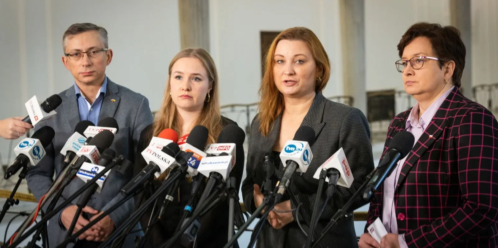 Cztery osoby występują na konferencji prasowej, stojąc przed rzędem mikrofonów z logo różnych mediów.