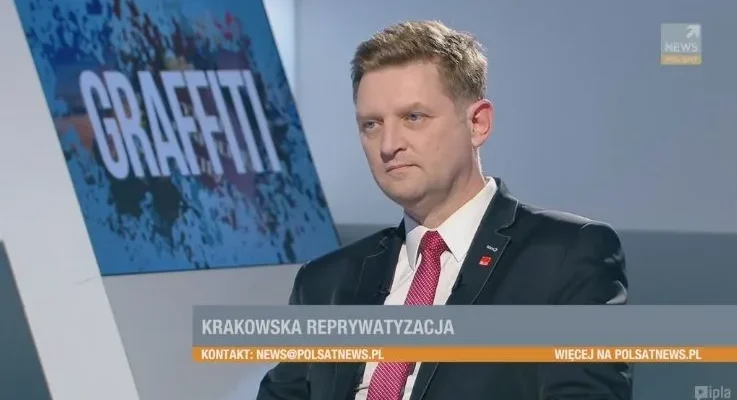 Mężczyzna w garniturze i czerwonym krawacie siedzi w studio telewizyjnym obok grafiki z napisem "GRAFFITI" i paskiem informacyjnym "KRAKOWSKA REPRYWATYZACJA".