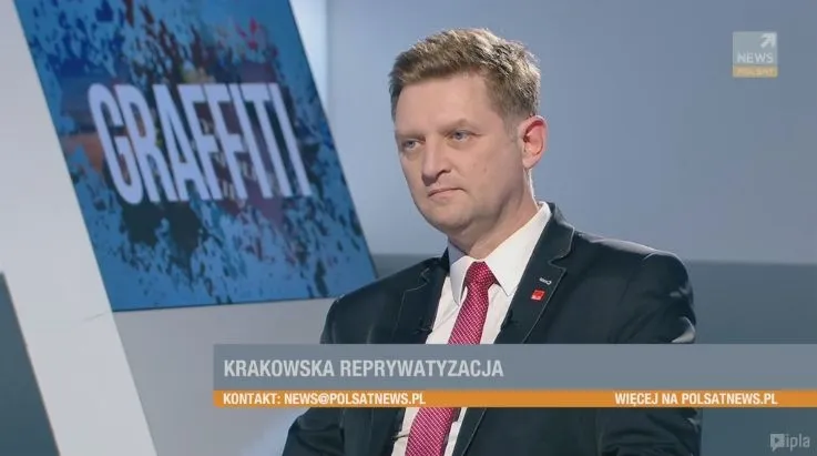 Mężczyzna w garniturze i czerwonym krawacie siedzi w studio telewizyjnym obok grafiki z napisem "GRAFFITI" i paskiem informacyjnym "KRAKOWSKA REPRYWATYZACJA".