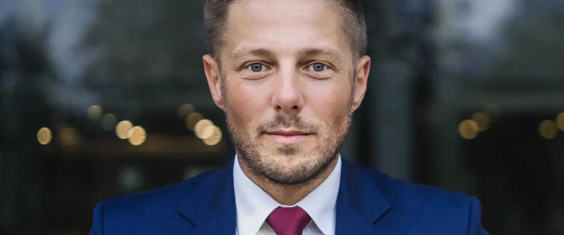 Portret mężczyzny w granatowym garniturze, białej koszuli i burgundowym krawacie, stojącego na tle rozmytych świateł.