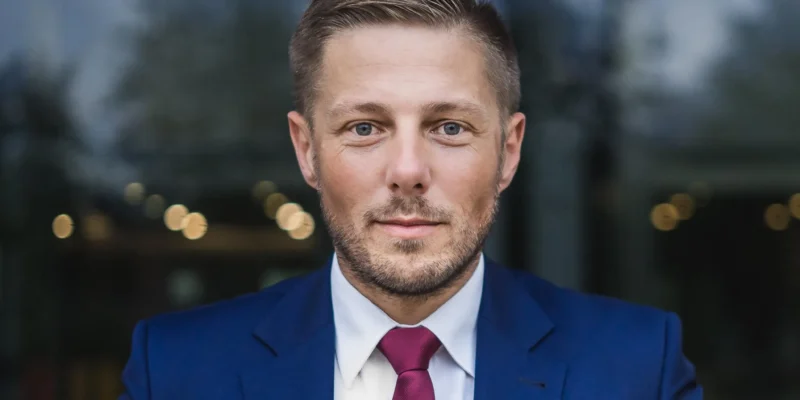 Portret mężczyzny w granatowym garniturze, białej koszuli i burgundowym krawacie, stojącego na tle rozmytych świateł.