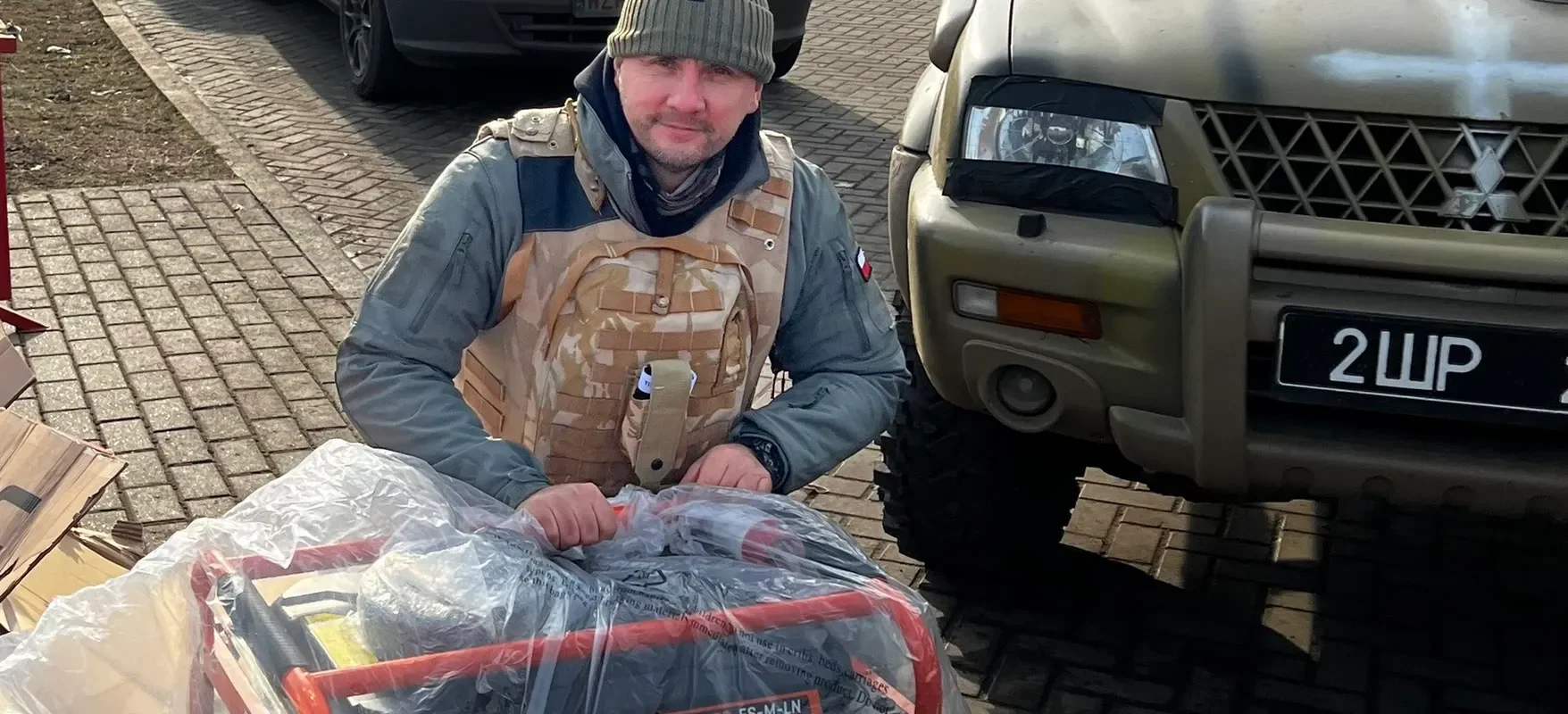 Mężczyzna w kamizelce ochronnej siedzi na opakowaniach obok generatora w otoczeniu samochodów zaparkowanych na ulicy.