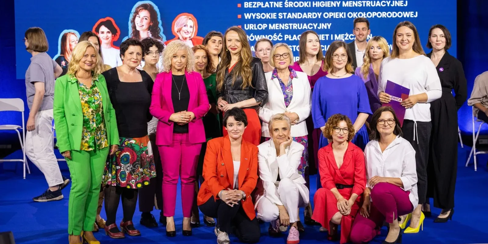 Grupa kobiet w różnobarwnych outfitach stoi i klęka na niebieskiej scenie podczas Konwencji Bezpieczna Polska, na tle dużego ekranu z listą punktów programu.