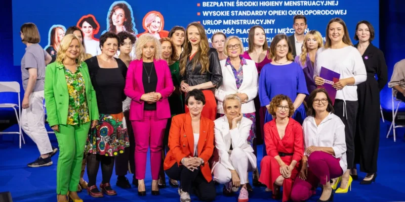 Grupa kobiet w różnobarwnych outfitach stoi i klęka na niebieskiej scenie podczas Konwencji Bezpieczna Polska, na tle dużego ekranu z listą punktów programu.