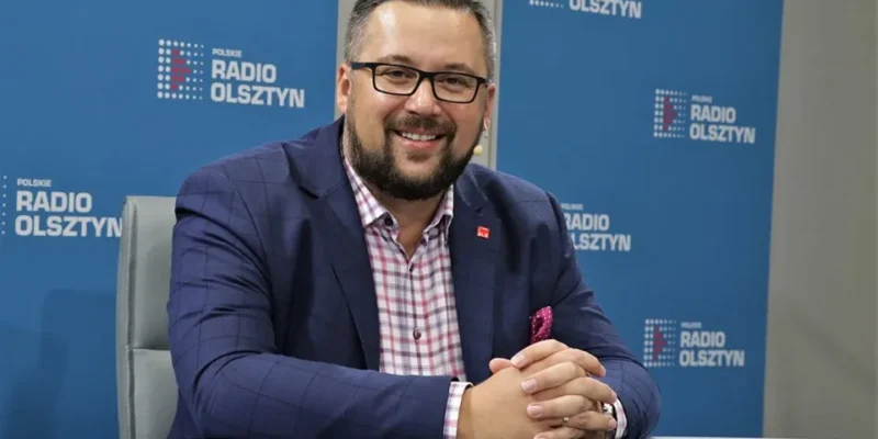 Mężczyzna w granatowym garniturze i różowej poszetce siedzi przy biurku w studiu Polskiego Radia Olsztyn i uśmiecha się do kamery.