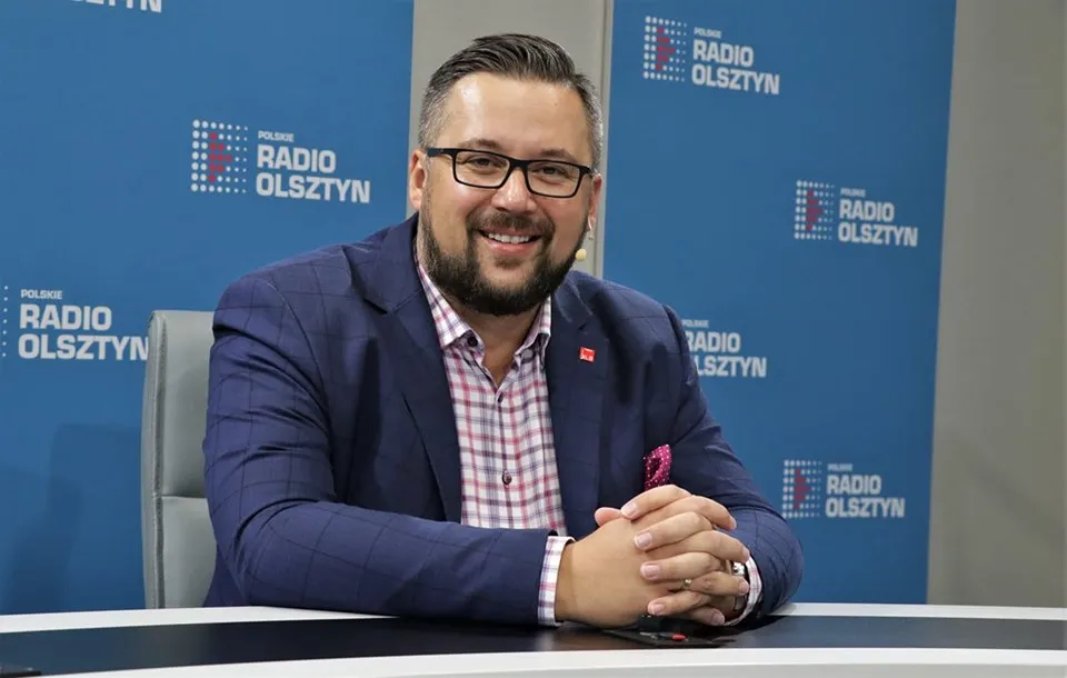 Mężczyzna w granatowym garniturze i różowej poszetce siedzi przy biurku w studiu Polskiego Radia Olsztyn i uśmiecha się do kamery.