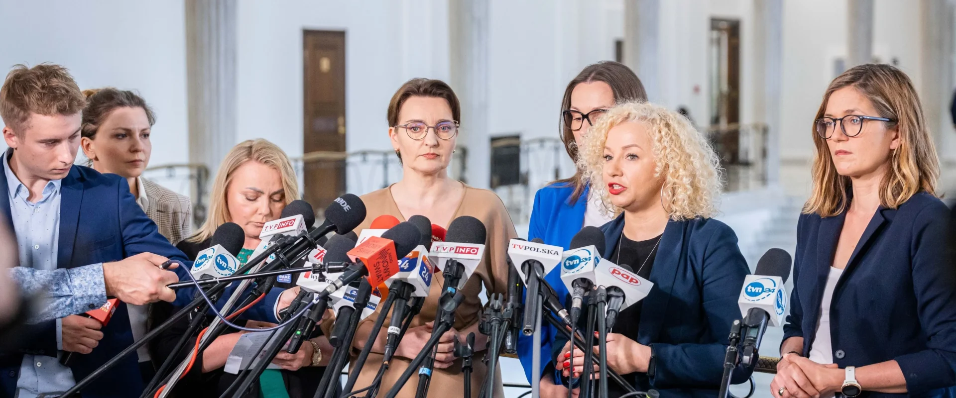 Na zdjęciu przedstawiono grupę polityków w trakcie konferencji prasowej, otoczoną mikrofonami i dziennikarzami.