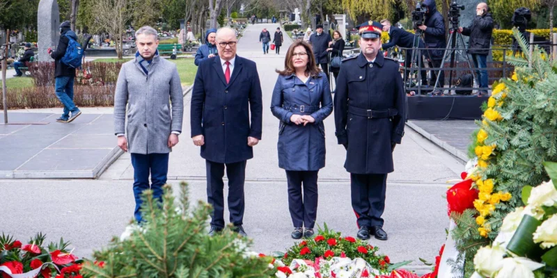 Czterech ludzi stoi w parku otoczonych kwiatami i wieńcami upamiętniającymi 12. rocznicę tragedii smoleńskiej, przy obecności kamer i fotoreporterów w tle.