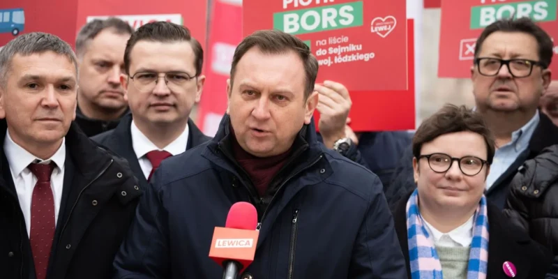 Mężczyzna w ciemnej kurtce przemawia do kamery na wiecu Lewicy, otoczony innymi uczestnikami i czerwonymi plakatami w tle.