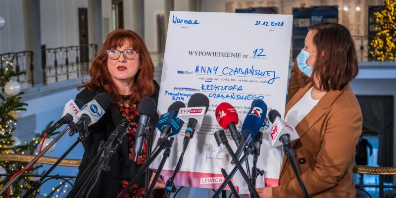 Na zdjęciu dwie kobiety prezentują dokument podczas konferencji prasowej, otoczone mikrofonami, w tle widoczna choinka.