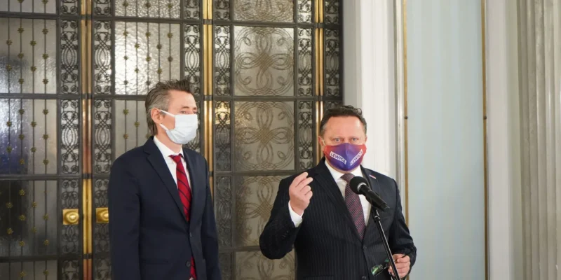 Dwóch liderów Lewicy i SPD w garniturach i maseczkach stoją przy mikrofonie w eleganckim wnętrzu, na tle ozdobnej metalowej bramy w tle.