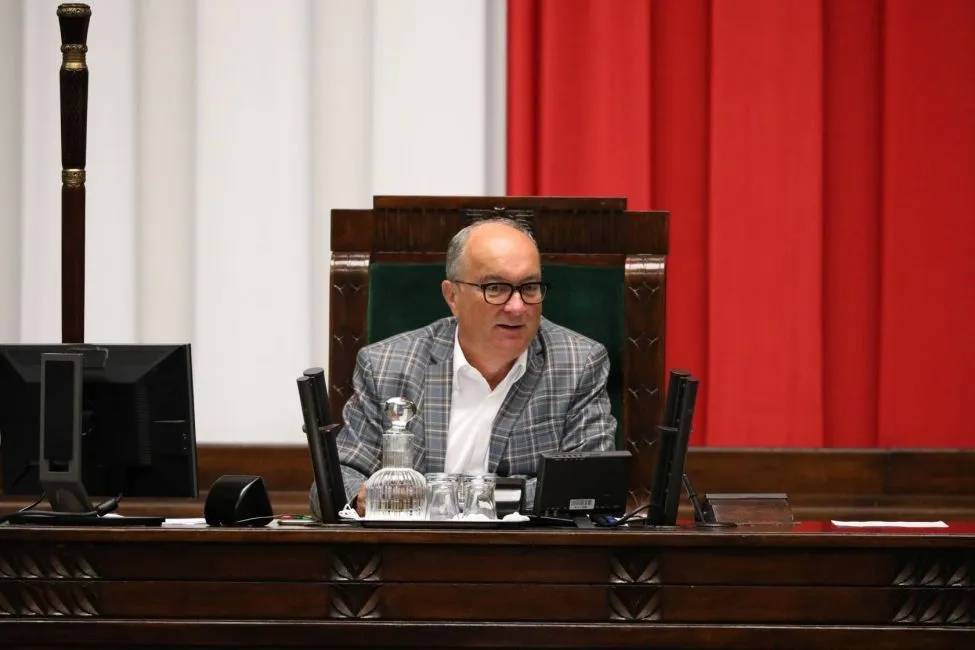 Polityk w marynarce w kratę siedzi za biurkiem w sali plenarnej, przed nim mikrofony i dzbanek z wodą, a w tle czerwone zasłony.