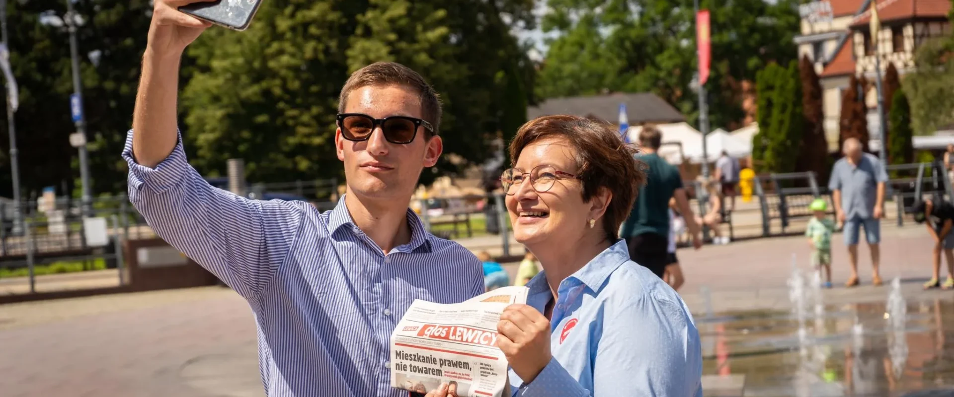 Mężczyzna w okularach przeciwsłonecznych robi selfie z kobieta trzymającą gazetę „Głos Lewicy” na placu z fontanną w tle.