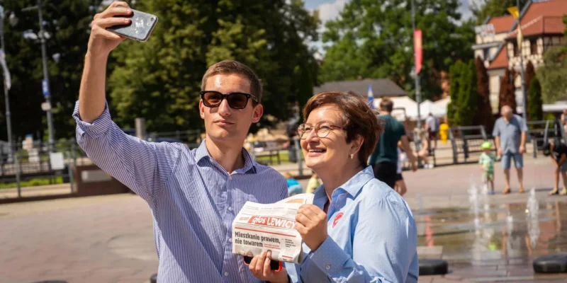 Mężczyzna w okularach przeciwsłonecznych robi selfie z kobieta trzymającą gazetę „Głos Lewicy” na placu z fontanną w tle.
