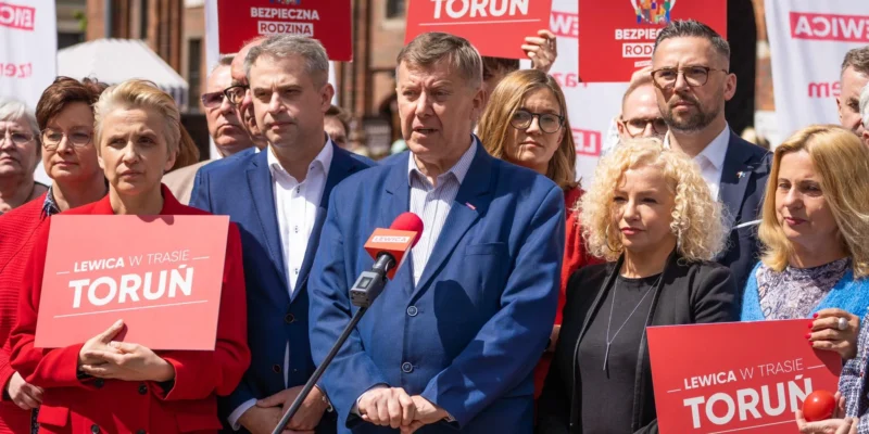 Grupa osób w formalnych ubraniach stoi na konferencji prasowej, trzyma plakaty z napisem "Lewica w trasie Toruń" i przemawia do mikrofonu.