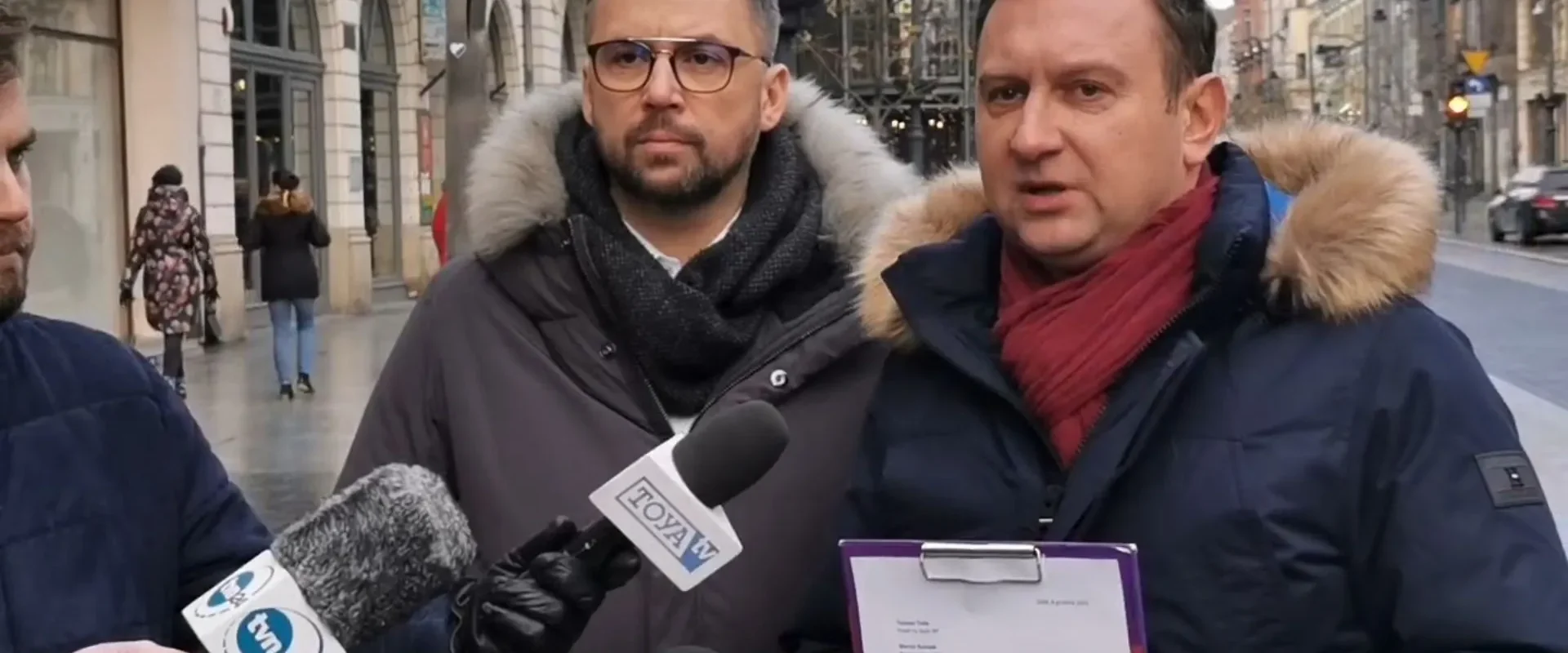 Dwóch mężczyzn w zimowych kurtkach z futrzanymi kapturami udziela wywiadu mediom na miejskim deptaku podczas interwencji Lewicy; jeden z nich trzyma teczkę z dokumentami.