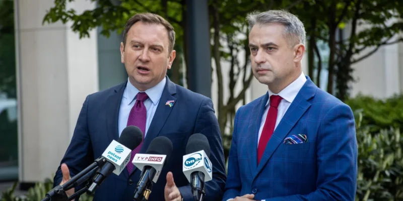 Dwaj mężczyźni stojący przy mikrofonach podczas konferencji prasowej, w tle zielone rośliny.