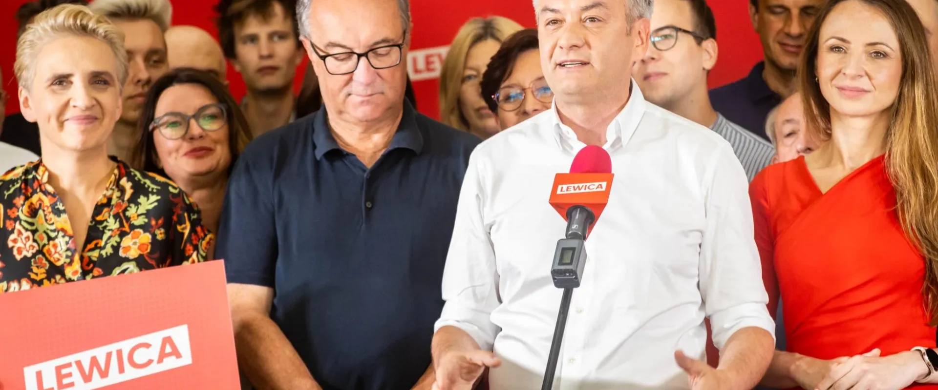 Grupa polityków Lewicy podczas konferencji prasowej; mężczyzna w białej koszuli przemawia do mikrofonu z logiem Lewica, otoczony współpracownikami i zwolennikami na czerwonej ścianie z banerami Lewicy.