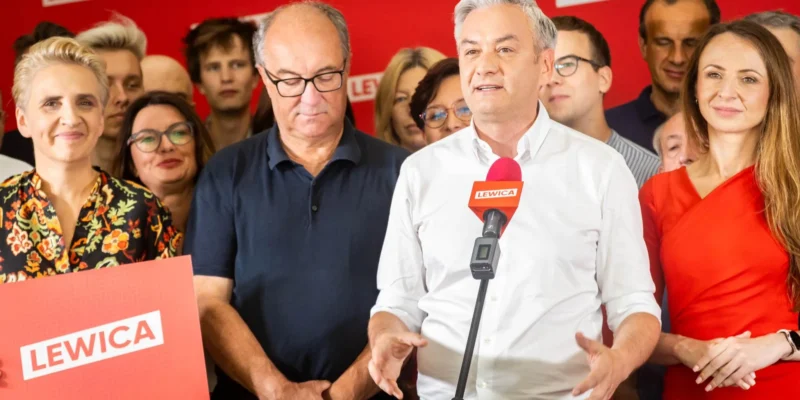 Grupa polityków Lewicy podczas konferencji prasowej; mężczyzna w białej koszuli przemawia do mikrofonu z logiem Lewica, otoczony współpracownikami i zwolennikami na czerwonej ścianie z banerami Lewicy.