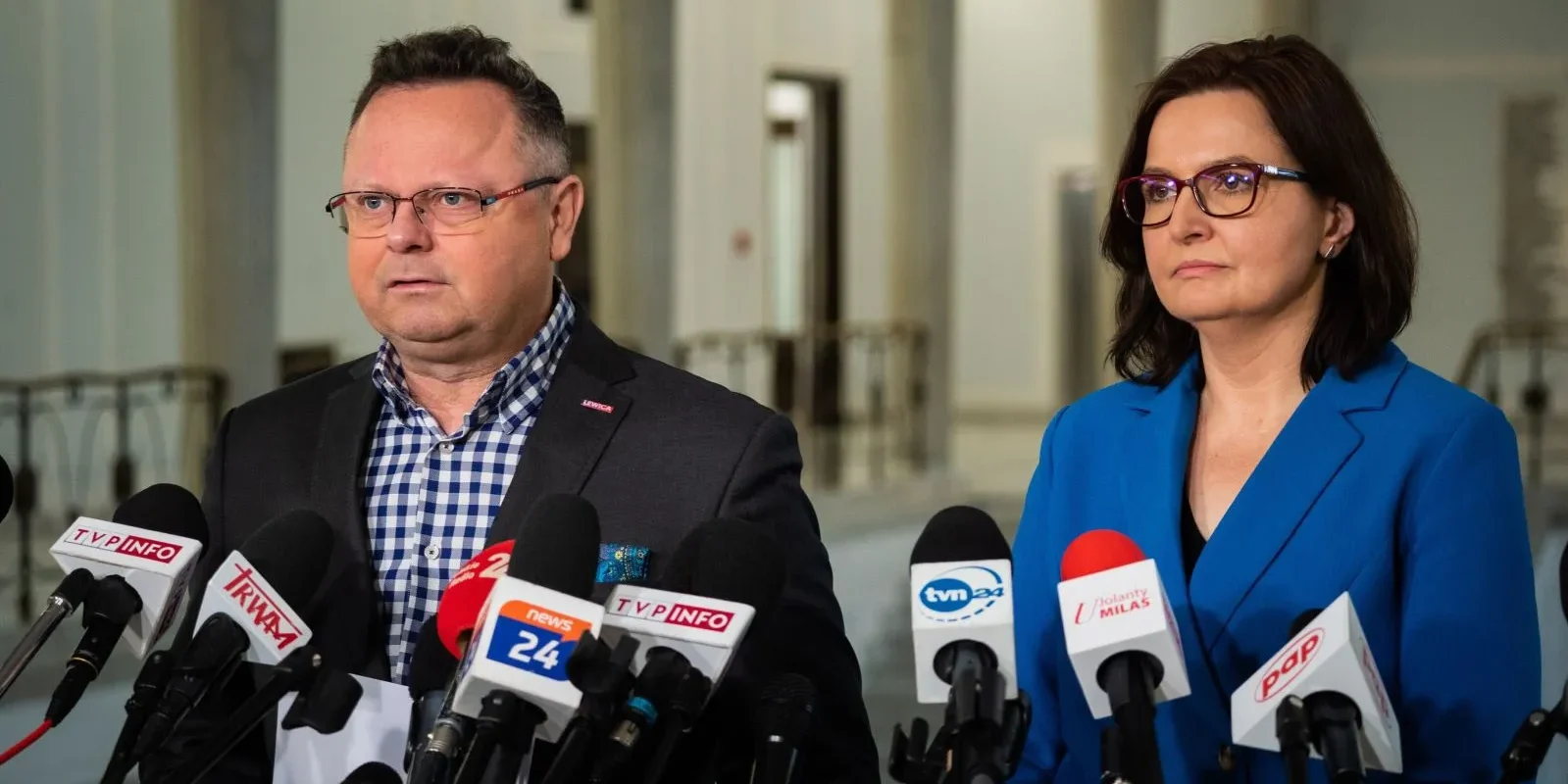 Dwoje dorosłych w garniturach stoi przed licznymi mikrofonami reporterów podczas konferencji prasowej.