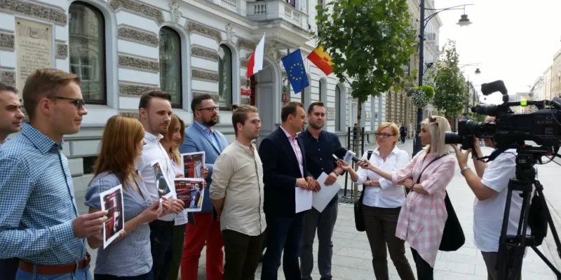 Grupa młodych osób stoi przed reprezentacyjną kamienicą z flagami, podczas gdy przedstawiciel MS prezentuje „4‑pak studencki” i udziela wywiadu dziennikarzom z mikrofonami i kamerą.