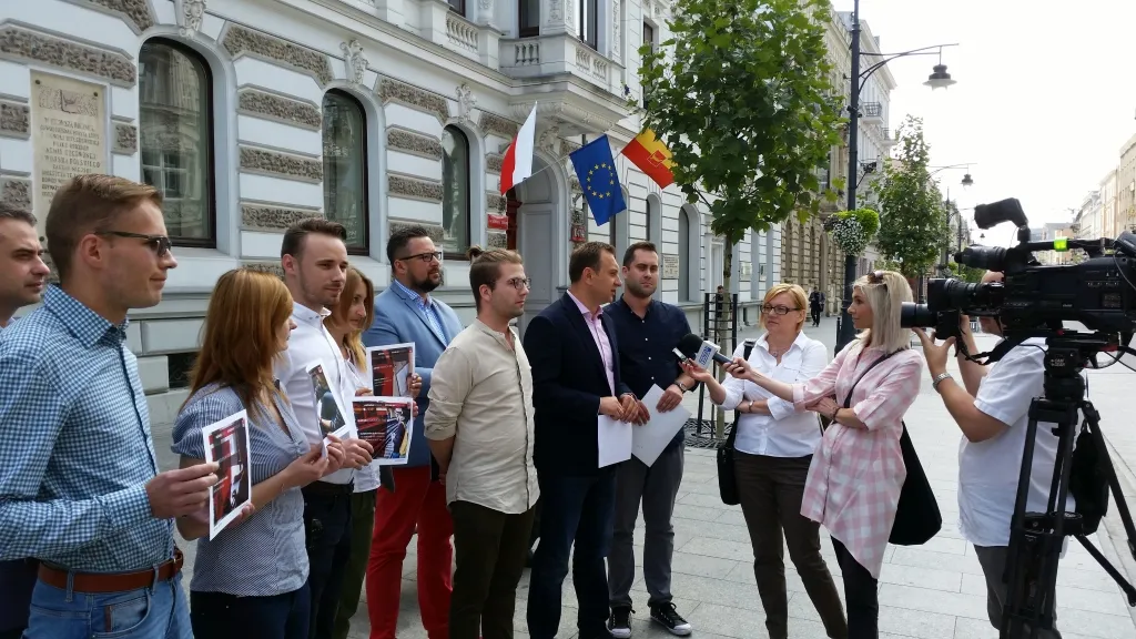 Grupa młodych osób stoi przed reprezentacyjną kamienicą z flagami, podczas gdy przedstawiciel MS prezentuje „4‑pak studencki” i udziela wywiadu dziennikarzom z mikrofonami i kamerą.